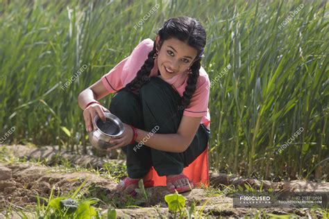 Happy Indian Girl Watering Small Plants Sitting In Agriculture Field