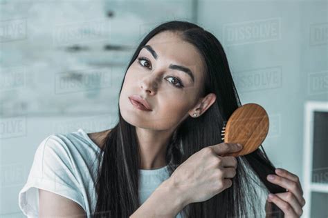 Young Brunette Woman Combing Hair With Hairbrush And Looking At Camera