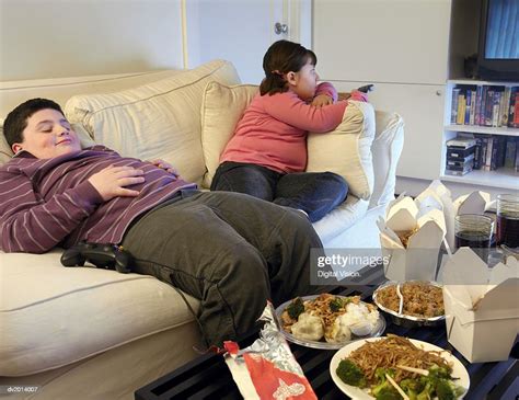 Overweight Brother And Sister Sitting On A Sofa Eating Takeaway Food