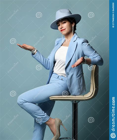 Smiling Short Haired Brunette Woman In Blue Business Suit And Hat Sits On Stool Holding Hand Up