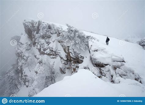 Man Walks Along The Cliff To The Top In The Snowy Misty Mountains