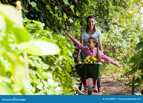 Smiling Woman Pushing Girl Sitting In Wheelbarrow On Footpath Amidst