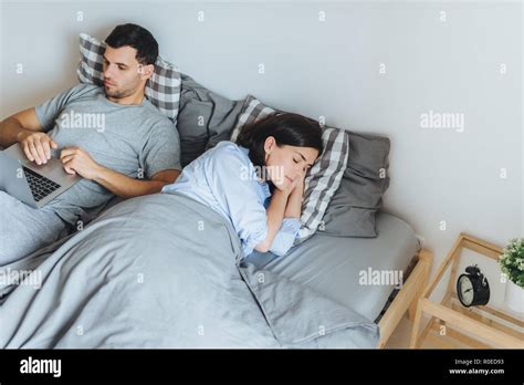 pretty female sleeps in bed sees pleasant dreams while her husband works on laptop computer