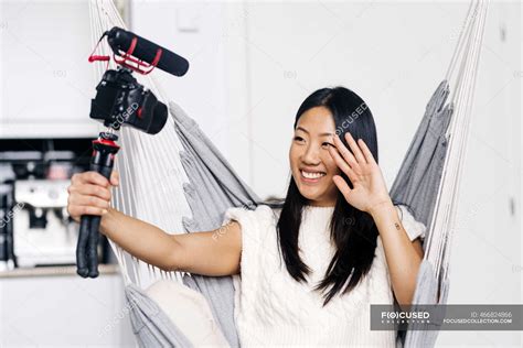 Smiling Ethnic Female Vlogger Recording Video On Photo Camera While Sitting On Hammock In The