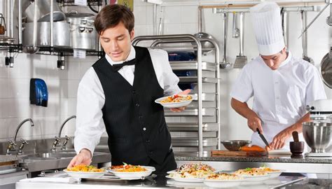 Young Waiter Placing Dishes In Tray With Chef Working In Commercial