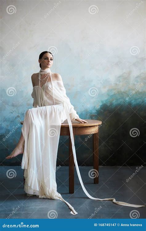 Beautiful Slender Brunette Girl Sitting On The Sofa In A Long White