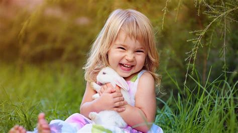 Smiley Cute Baby Girl Is Sitting On Green Grass Holding Rabbit Wearing