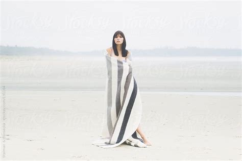 A Woman Standing On Top Of A Beach Covered In A Blanket By An Ocean Shore