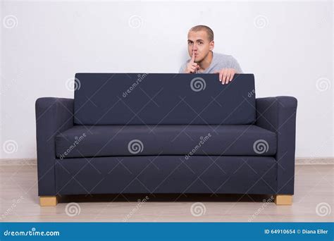 Young Man Hiding Behind A Sofa And Showing Shhh Sign Stock Photo