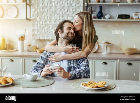 Happy Caucasian Millennial Blonde Wife Kissing Man With Stubble On Light Kitchen Interior Sun