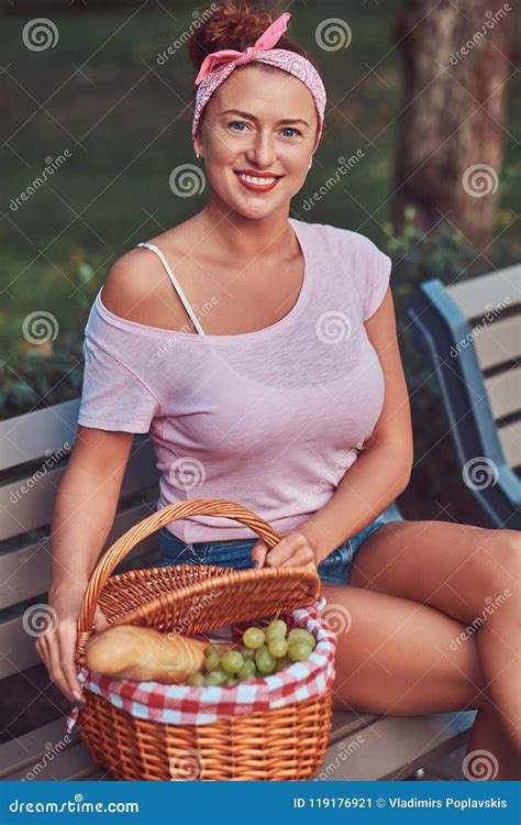 Happy Beautiful Redhead Female Wearing Casual Clothes Sitting With A Picnic Basket On A Bench In
