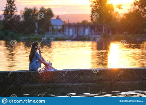 Back View Of Lonely Woman Sitting On Lake Shore On Warm Evening
