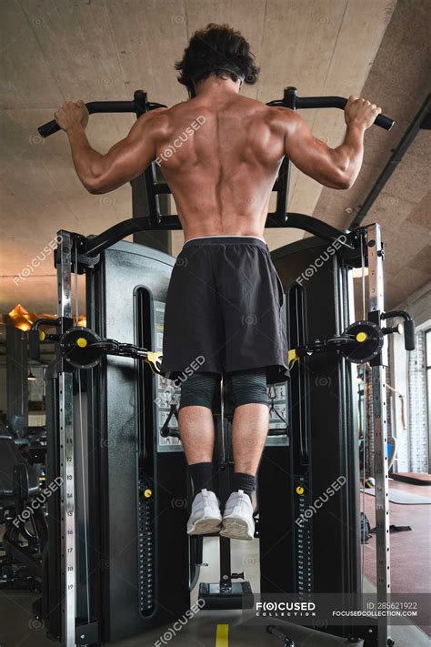 Back View Of Shirtless Man Performing Pull Ups On Exercise Machine