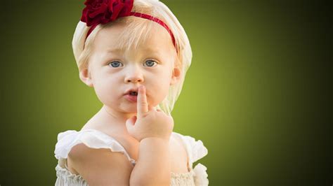 Cute Little Girl Baby Is Keeping Finger On Lips In Green Blur Background Wearing White Dress And ...