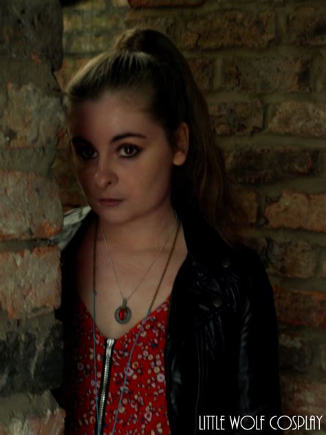 a woman standing in front of a brick wall wearing a black leather jacket and necklace