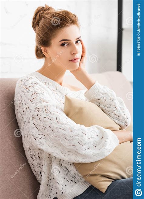 Woman In White Sweater Holding Pillow And Looking At Camera Stock Photo
