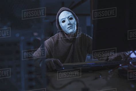 Computer Hacker Wearing Mask And Hood Using Computer While Sitting At