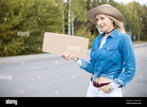 Modern Grimacing Sarcastic Blond Woman In Hat Hitchhiking With Blank Carton Board And Wait For