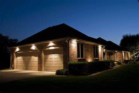 Two Garage Doors Are Lit Up At Night In Front Of A House With Bushes