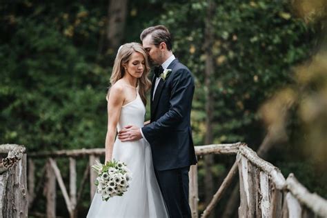 A Bride And Groom Standing On A Wooden Bridge In The Woods With Their Arms Around Each Other