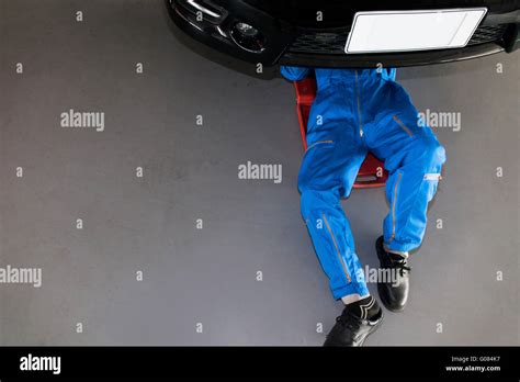 Mechanic In Blue Uniform Lying Down And Working Under Car At The Garage