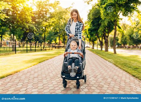 Young Mother Walks With A Little Toddler In A Stroller In The Summer On