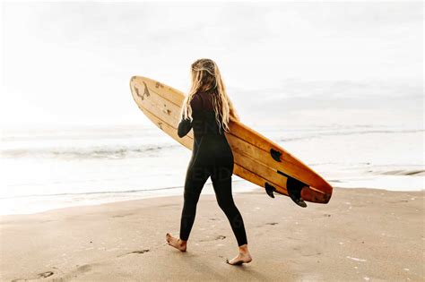 Side View Of Surfer Woman Dressed In Wetsuit Walking While Carrying Surfboard Above Head On The