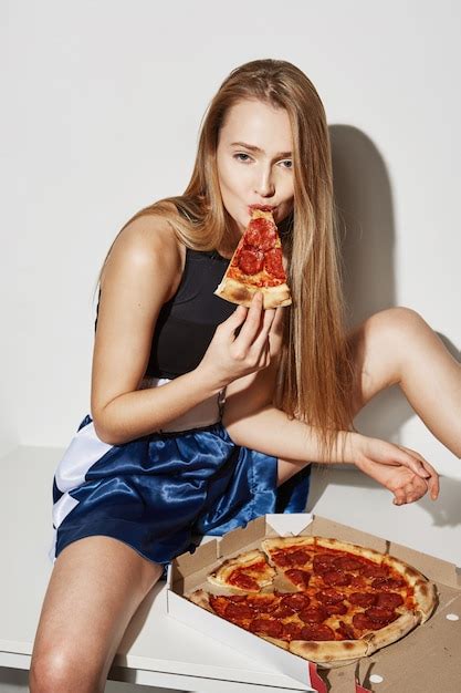 frau mit blonden haaren sitzt mit gespreizten beinen auf dem tisch und isst pizza mit flirtendem