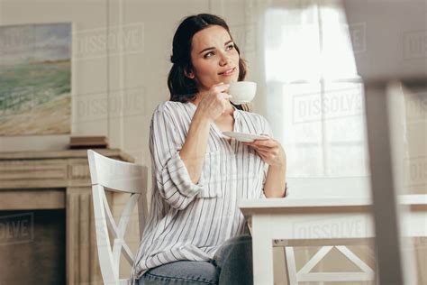 Pensive Young Woman Drinking Coffee While Sitting At Table At Home