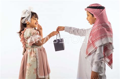 خلفية بيضاء لصبي وفتاه صغيران سعوديان مبتسمان ، يعطي الطفل السعودي هدية بكيس أسود لطفلة سعودية