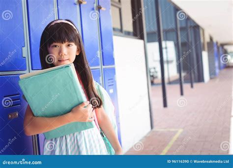 Portrait Of Elementary Girl Holding Books In Corridor Stock Photo