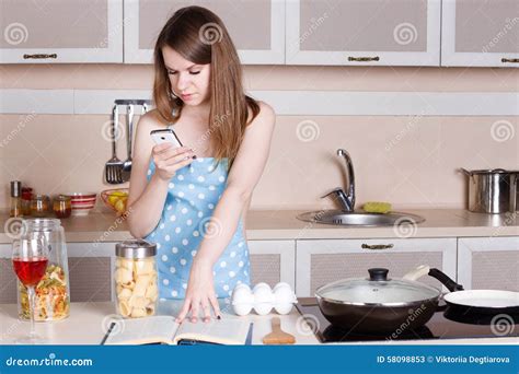 Girl In The Kitchen Wearing An Apron Over His Naked Body Prepares And Looks Into Phone Stock