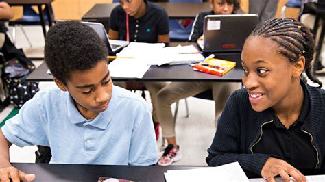 A Smiling Girl Looks At A Boy Who Is Reviewing A Paper On A School Desk