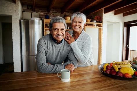 Portrait Of Happily Retired Elderly Biracial Couple Leaning On Kitchen Counter Smiling At
