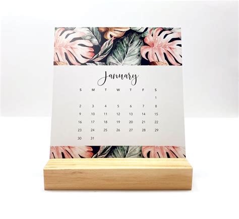 A Desk Calendar Sitting On Top Of A Wooden Stand With Tropical Leaves