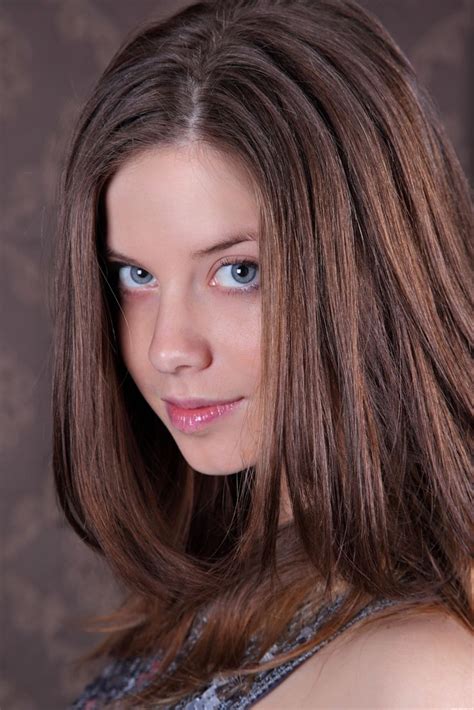 A Woman With Long Brown Hair And Blue Eyes Posing For A Photo In Front Of A Wall