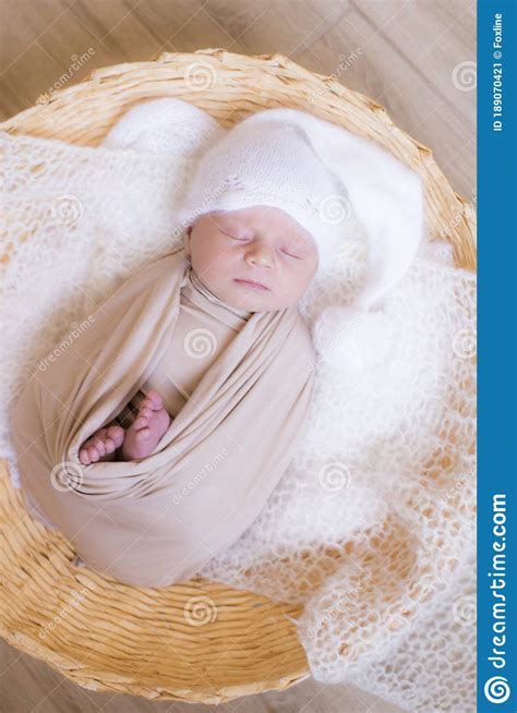 Cute Little Baby In White Knitted Hat Lies In A Wicker Basket In A