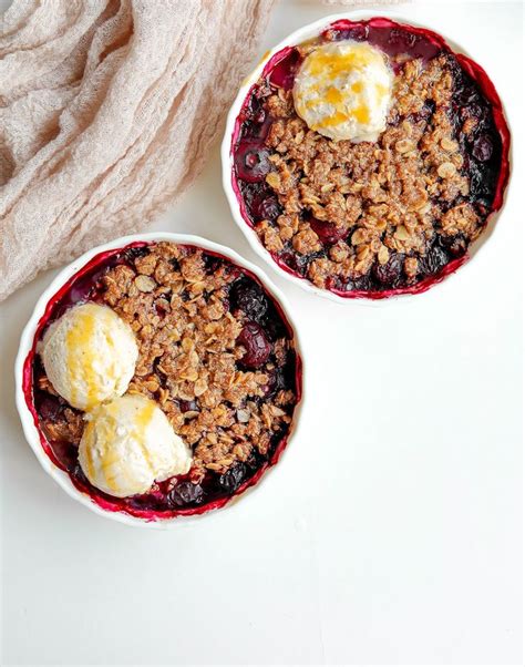 Two Bowls Filled With Blueberry Crumble And Ice Cream On Top Of A White