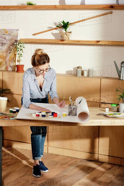 Stylish Young Female Artist In Eyeglasses Artist Painting At Table In