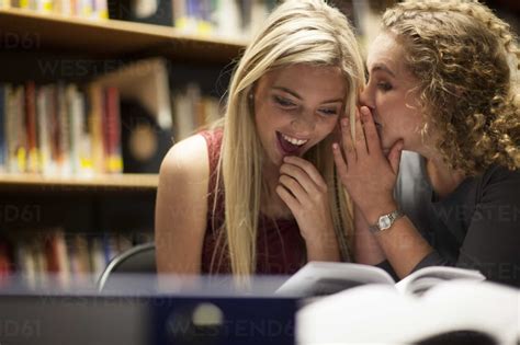 Two Female Students Whispering In A Library Stock Photo