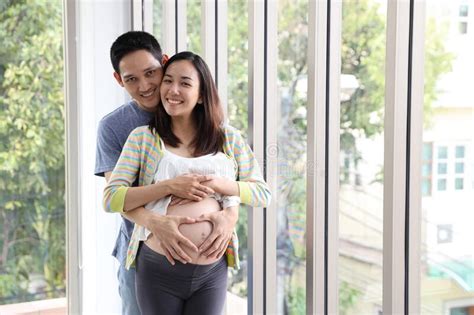 Asian Husband Giving Kiss To Young Pregnant Belly Wife With Love On Sofa In Living Room Stock