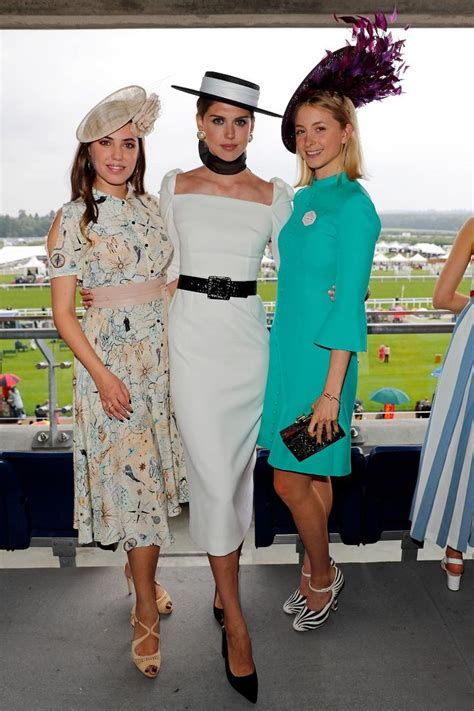 Three Women Standing Next To Each Other In Dresses And Hats At A Horse