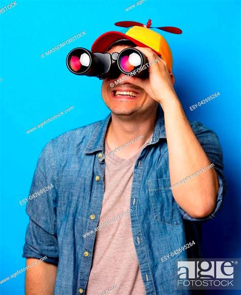 Young Nerd Man With Noob Hat Using A Binoculars On Blue Background