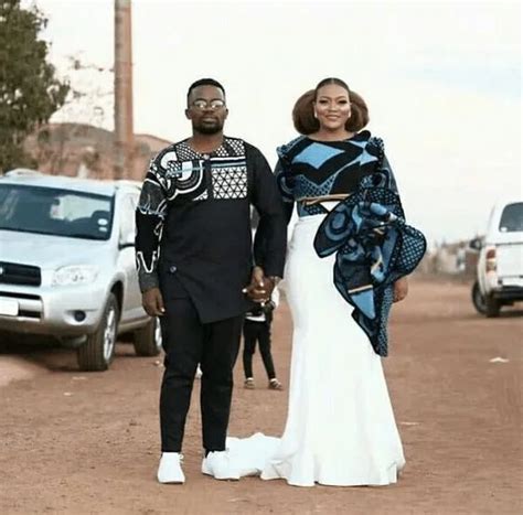 A Man And Woman Standing In The Middle Of A Dirt Road With Cars Behind Them