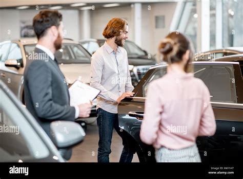 Young Couple With Salesperson Choosing A New Car Looking On The Back