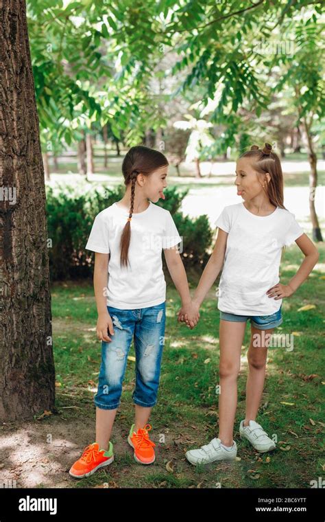 Two Little Girls In White T Shirts Stand Holding Hands In Park Outdoor