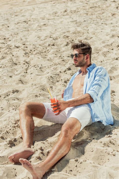 Handsome Bearded Man In Sunglasses Sitting On Sandy Beach With Glass Of