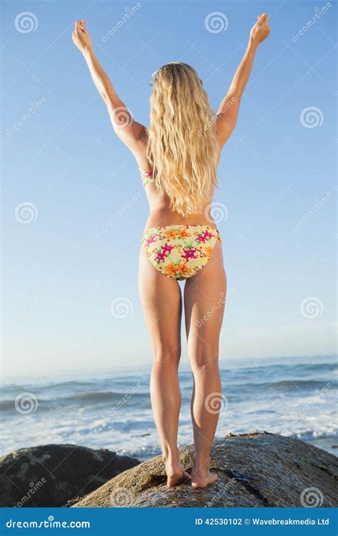 Herrliche Blondine In Anhebenden Armen Des Blumenbikinis Auf Einem Felsen Am Strand Stockfoto