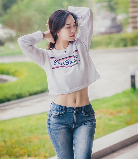 A Woman In Jeans And A Crop Top Is Standing On The Sidewalk With Her Hands Behind Her Head