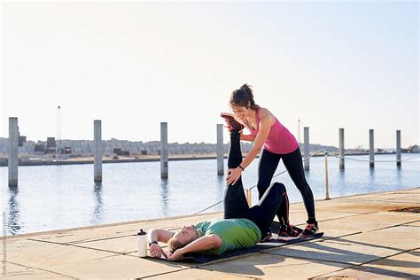 Woman Helping Man To Stretch Leg On Mat On Pier By Stocksy Contributor Guille Faingold